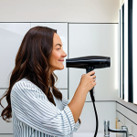 A professional adult woman, mid-30s, with a serene expression, efficiently drying her healthy, shiny hair in a bright, modern bathroom. She holds a sleek, lightweight hair dryer, showcasing its ergonomic design and comfortable grip. Her hair is smooth and frizz-free, illustrating the benefits of advanced hair care technology. She is wearing modest, comfortable home attire. The scene is well-lit, emphasizing a peaceful morning routine. Fully clothed, appropriate attire, safe for work, perfect anatomy, correct proportions, natural pose, well-formed hands, proper finger count, natural body proportions, professional, family-friendly, appropriate content.